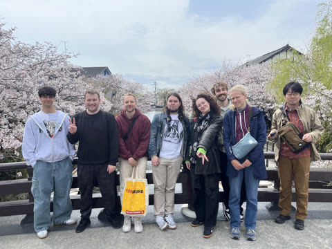 A group of eight people stands posing on a bridge over a river lined with blooming cherry blossom trees.