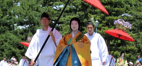 A person in an orange kimono walks between two attendants in white robes holding red parasols during an outdoor procession.