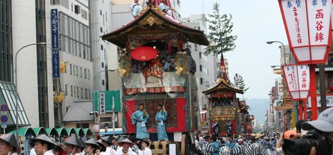 Large, ornate floats carrying people move in a procession down a city street lined with buildings.