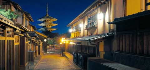 A multi-tiered pagoda stands over a narrow, stone-paved street lined with traditional, illuminated wooden buildings at night.