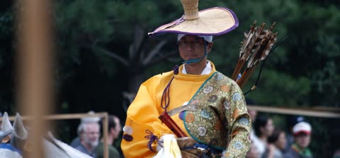 A person in a yellow and patterned robe, a straw hat, and a quiver of arrows sits on a horse outdoors.