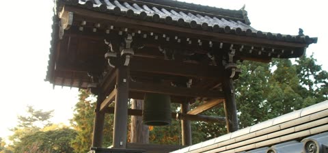 A large bell hangs within a wooden Japanese bell tower with a tiled roof, surrounded by green trees.