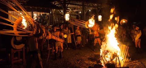 People carry large, flaming basket-like torches toward a central bonfire at an outdoor nighttime gathering.