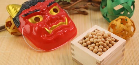 A red mask with golden fangs and a wooden box of soybeans sit beside woven balls on a bamboo mat.