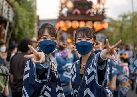 Two women in blue patterned jackets and face masks hold up peace signs during an outdoor festival.
