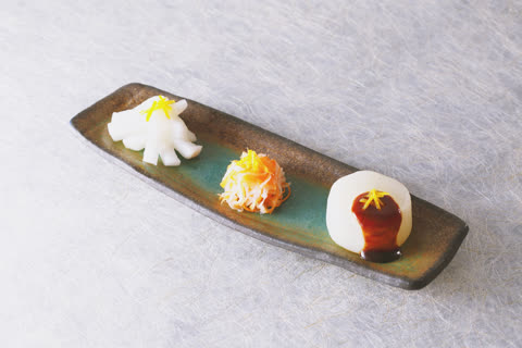 A long ceramic dish holds three small portions of garnished food: one flower-shaped, one shredded, and one round with sauce.