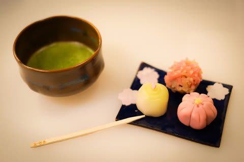 A brown ceramic bowl of matcha tea sits next to a blue plate holding three colorful wagashi sweets on a white surface.