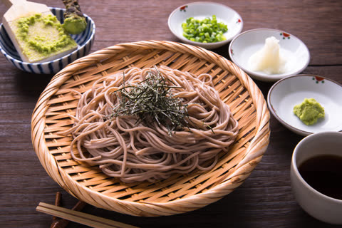A serving of soba noodles in a woven bamboo basket is arranged on a wood table with wasabi, green onions, and dipping sauce.