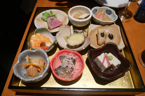 A collection of nine small Japanese dishes including beef, shrimp, and tofu are served on a gold and black tray.