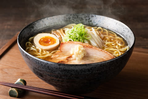 A steaming bowl of ramen with pork, a soft-boiled egg, and scallions sits on a wooden surface next to chopsticks.