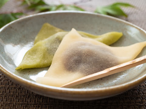 Two triangle-shaped Japanese sweets, one beige and one green, rest with a wooden pick in a ceramic bowl.