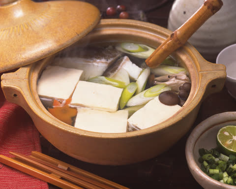 A steaming ceramic pot filled with fish, tofu, and vegetables in broth sits on a table next to chopsticks and garnishes.
