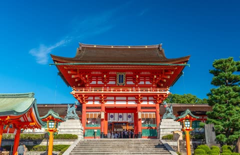 An ornate red Japanese building with a dark roof stands atop a stone staircase with people gathered at its entrance.