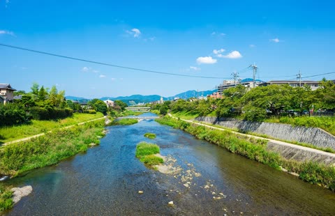 A shallow river flows between green banks through a small town, with mountains visible in the distance under a blue sky.