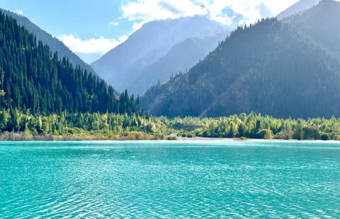 A bright turquoise lake sits at the base of steep mountains covered with dense evergreen forests under a partly cloudy sky.