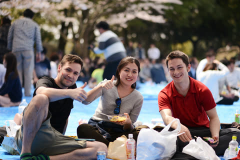 Two men and a woman sit on a blue tarp eating and gesturing thumbs-up towards the camera in a crowded park.