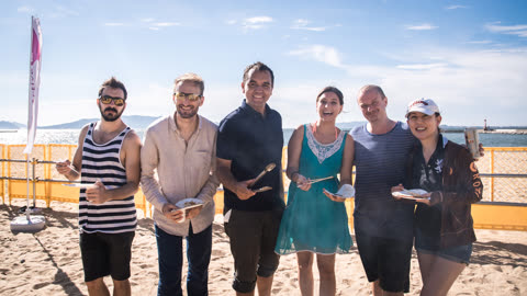 A group of six adults stands in a line holding plates of food on a sandy beach by the ocean.