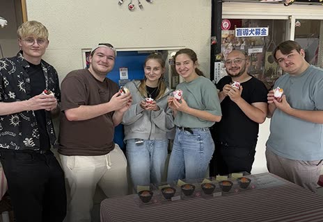 Six people stand together in a room, holding up small decorated desserts over a table with more desserts lined up.
