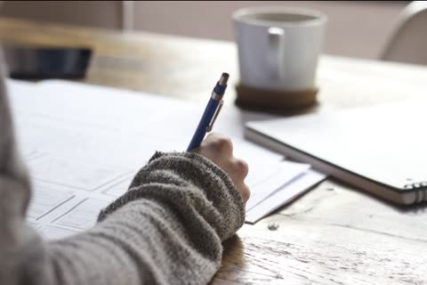 Person's hand writes with a blue pen on papers at a wooden desk.