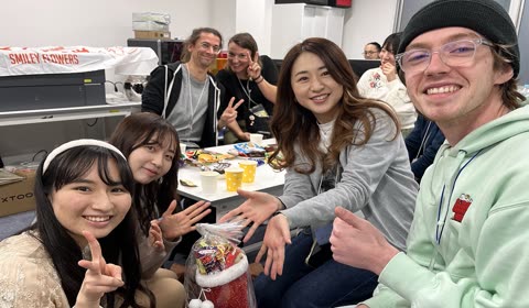 A diverse group of people smile and gesture toward the camera while gathered around a table in an office.