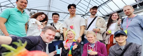 A group of ten smiling people poses for a photograph together inside a large greenhouse.