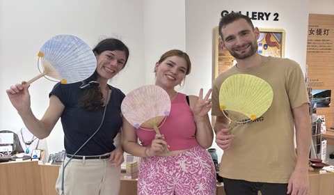 Two women and a man stand smiling while holding colorful, round paper fans inside an art gallery.