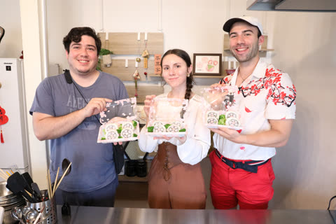 Two men and one woman smile while holding open containers of floral-patterned sushi rolls in a kitchen.