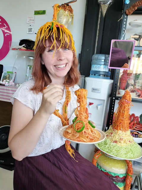 A smiling woman wears a fake spaghetti wig while holding a fork and a plate of fake spaghetti in a shop of food models.