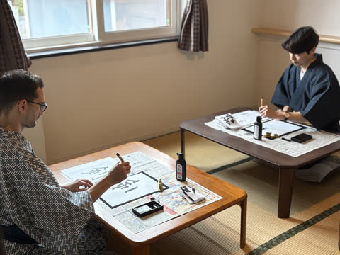 Two men in traditional robes practice calligraphy with brushes at low tables in a room with tatami mat floors.