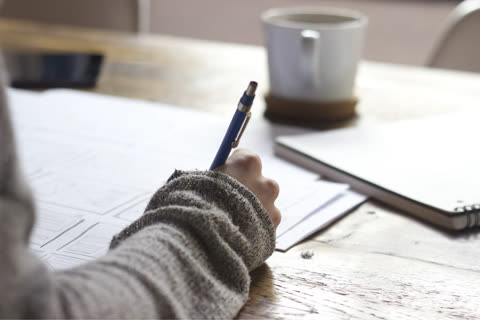 Person's hand writing with a blue pen on papers at a wooden desk.