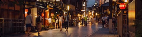People walk down a narrow street lined with traditional Japanese buildings and glowing lanterns at dusk.