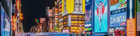Tall buildings covered in massive, bright neon signs with Japanese characters illuminate a crowded city canal at night.