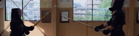 Two people in kendo armor holding bamboo swords face each other inside a dojo with large windows.