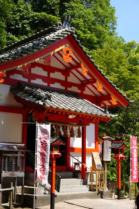 A red and white Japanese shrine with a grey-tiled roof stands on stone steps in front of tall green trees.