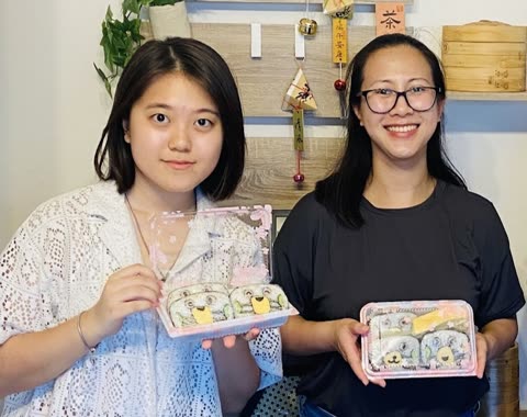 Two women stand side-by-side indoors, holding up clear containers of rice balls decorated to look like cartoon faces.