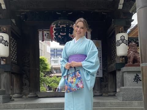 A woman in a light blue kimono smiles while holding a floral bag on the steps of a Japanese temple gate.