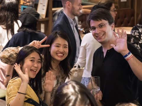 Two women and a man smile for a photo in a busy bar, with one woman making a peace sign and the others waving.