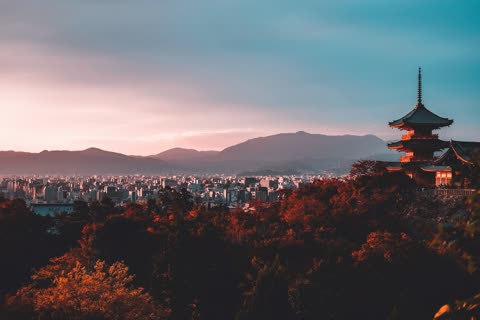 A multi-story red pagoda overlooks a dense city and mountain range from a hill at sunset.